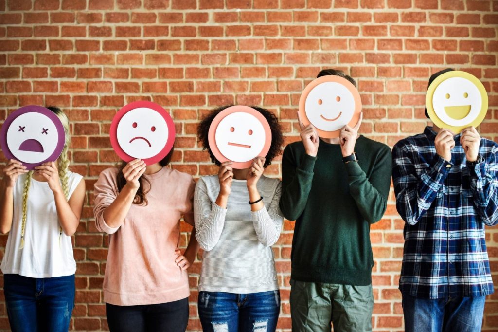 Five teenage boys and girls standing against a wall, holding a range of happy and sad emoji faces over their faces