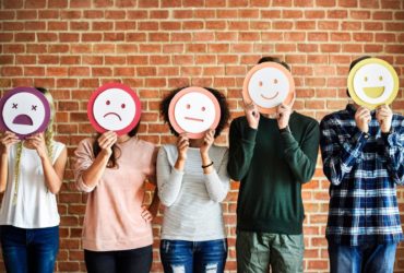 Five teenage boys and girls standing against a wall, holding a range of happy and sad emoji faces over their faces
