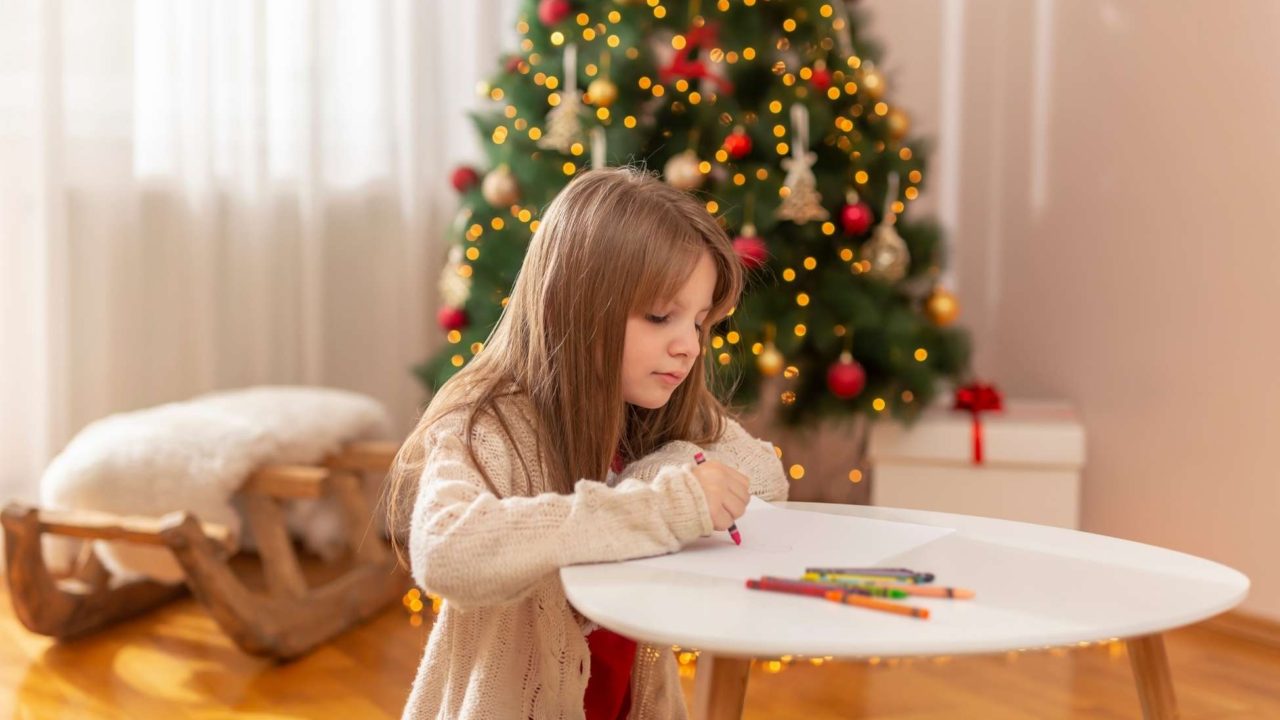 Girl drawing on a piece of paper with a Christmas tree behind her