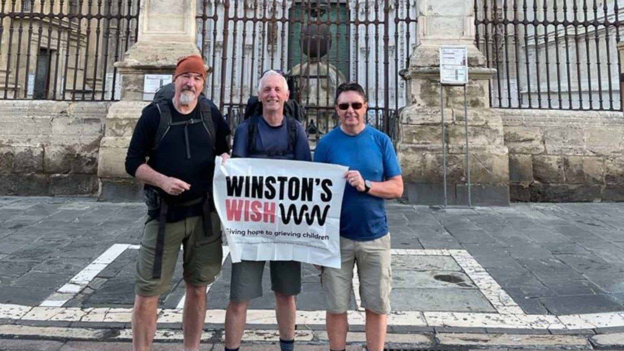 Barry, Alec and another man standing in front of a cathedral in Spain holding a Winston's Wish banner