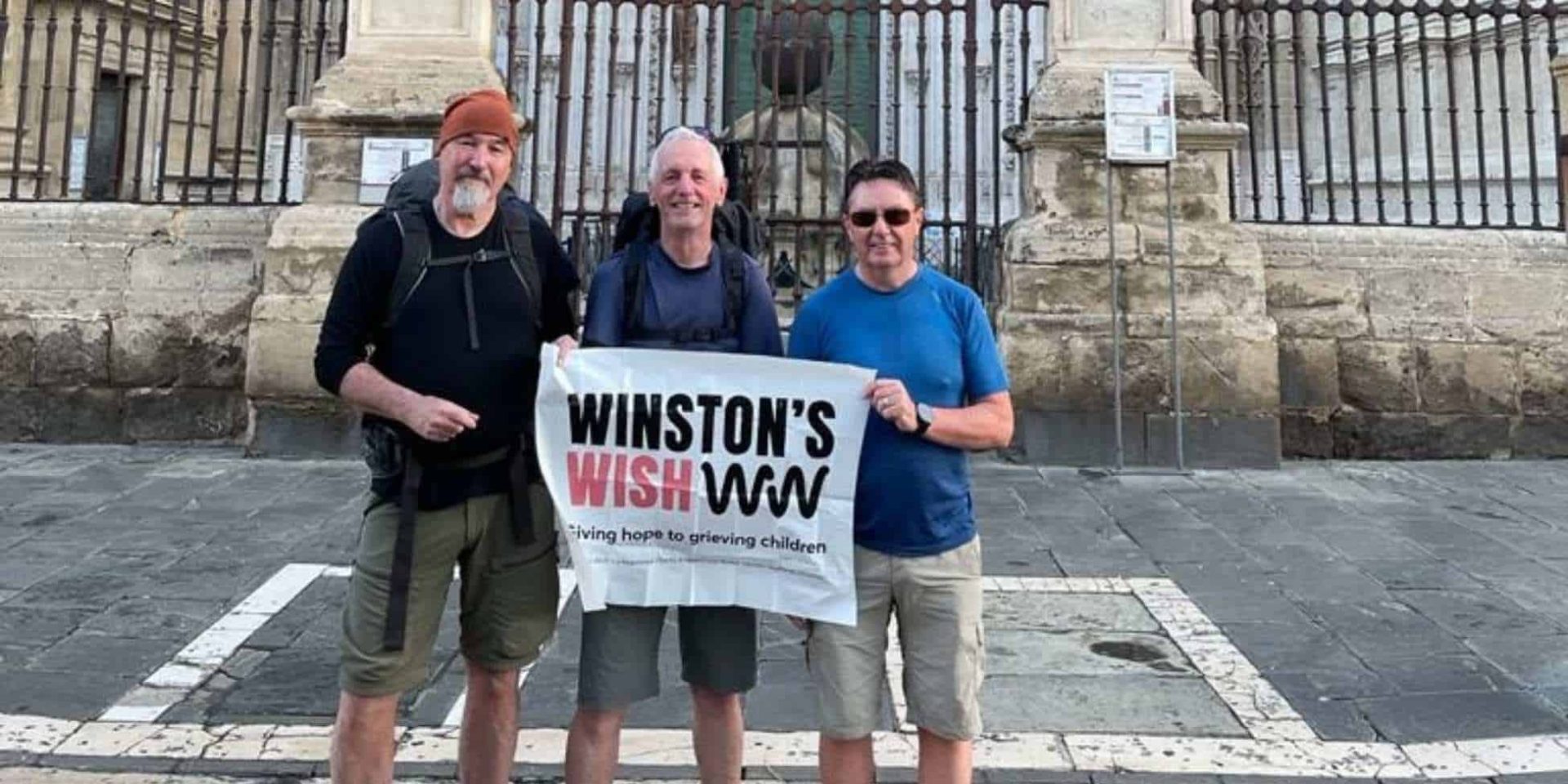 Barry, Alec and another man standing in front of a cathedral in Spain holding a Winston's Wish banner