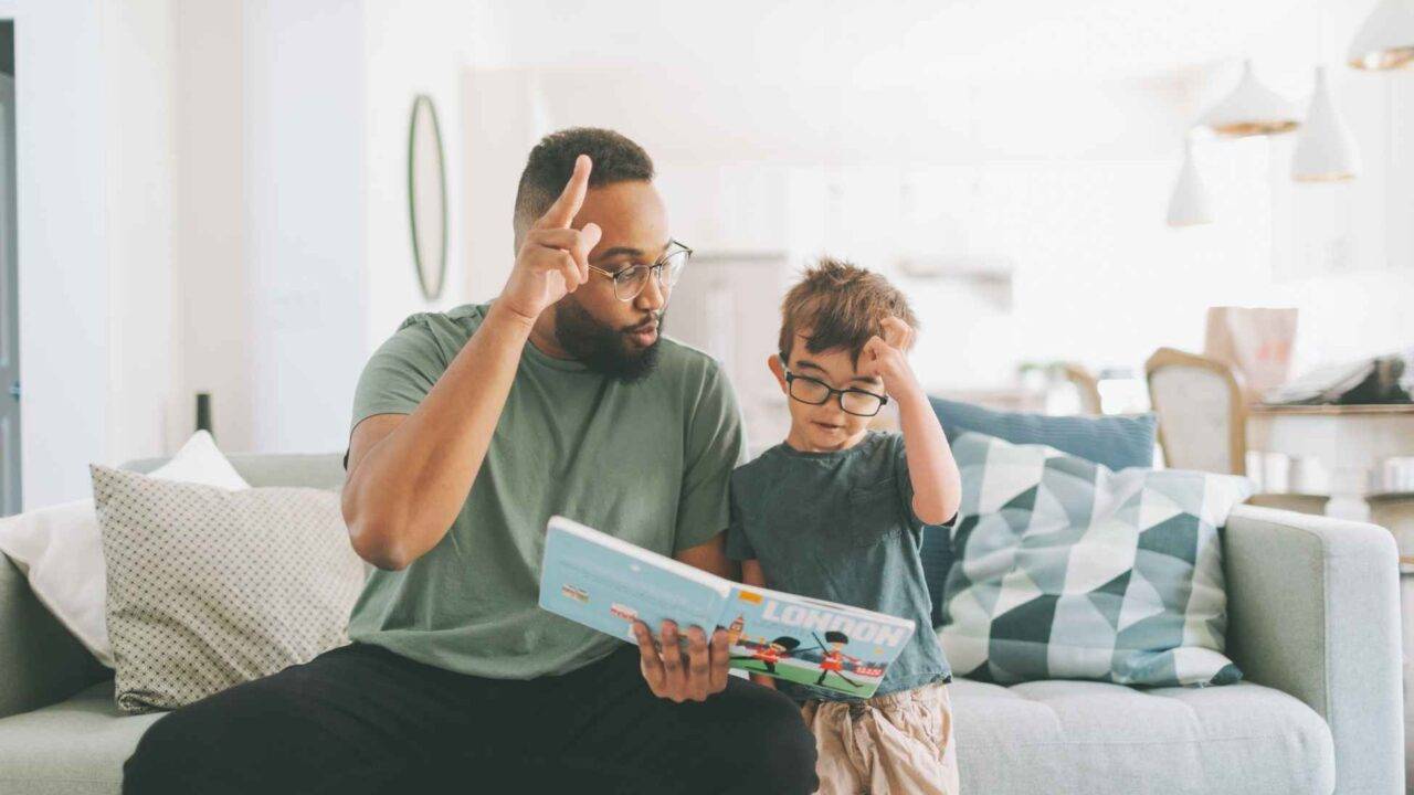 Dad and son sat on a sofa reading a book together and making hand gestures