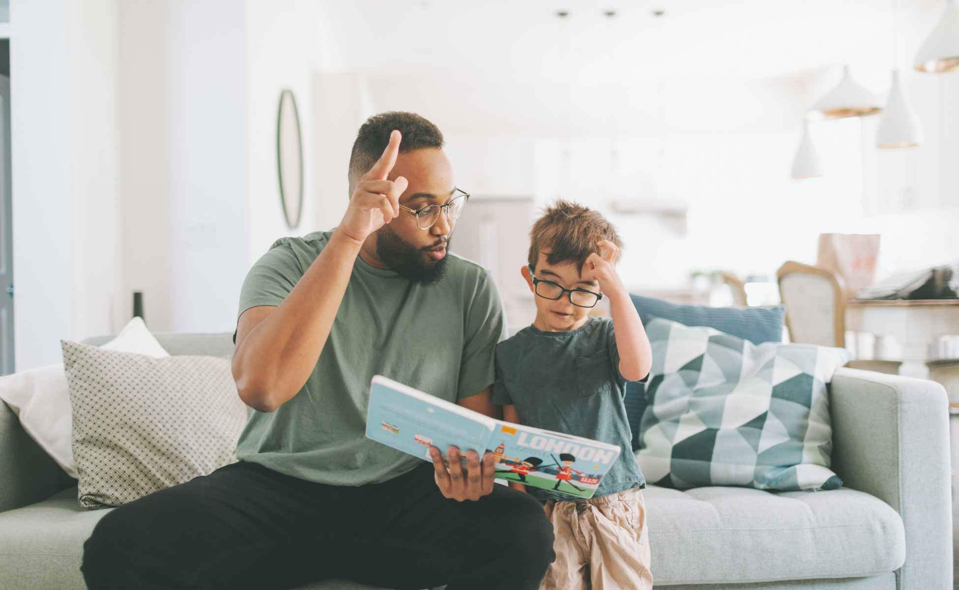 Dad and son sat on a sofa reading a book together and making hand gestures