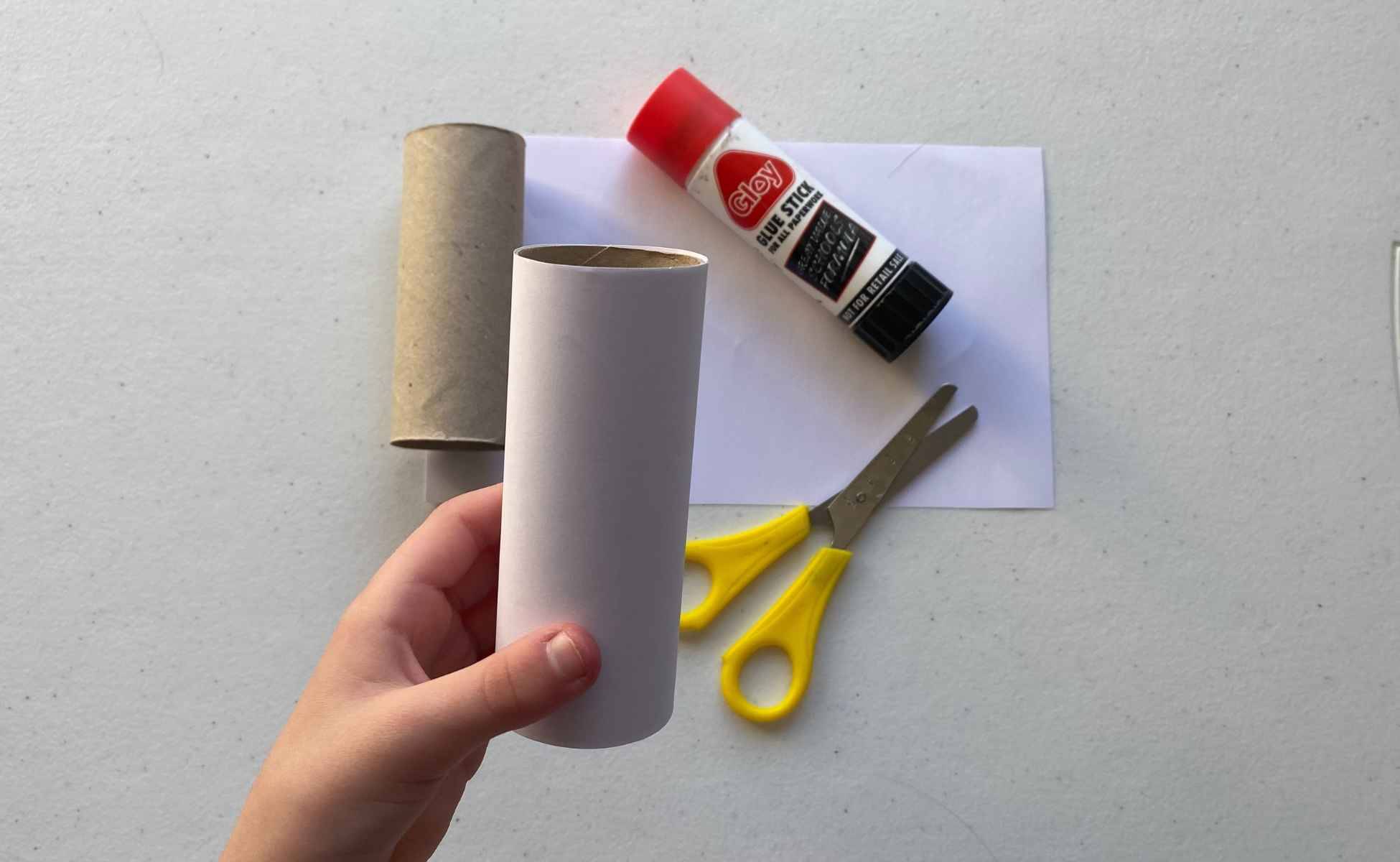 Child's hand holding toilet roll tube with glue, scissors and white paper on the table. Step one of making the snowman strike Christmas activity