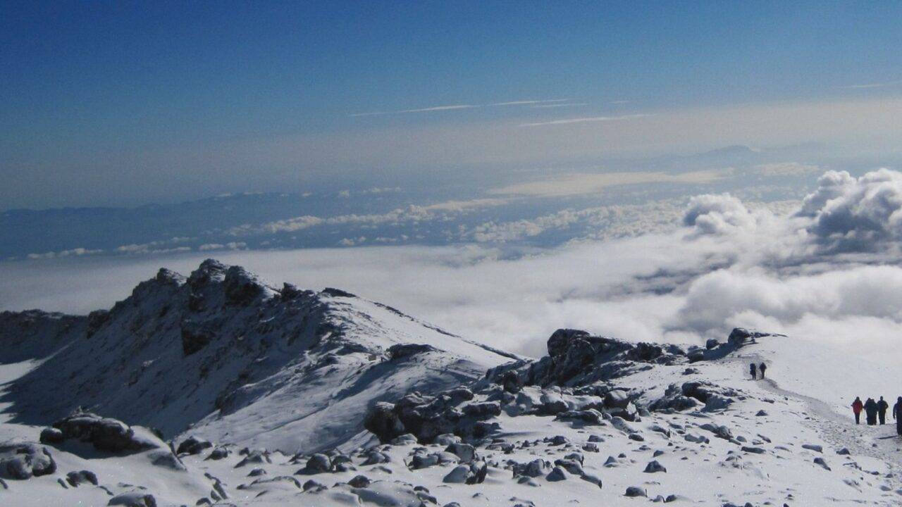 photograph of mount Kilimanjaro's summit with clouds and snow and blue sky, there are people walking to the peak