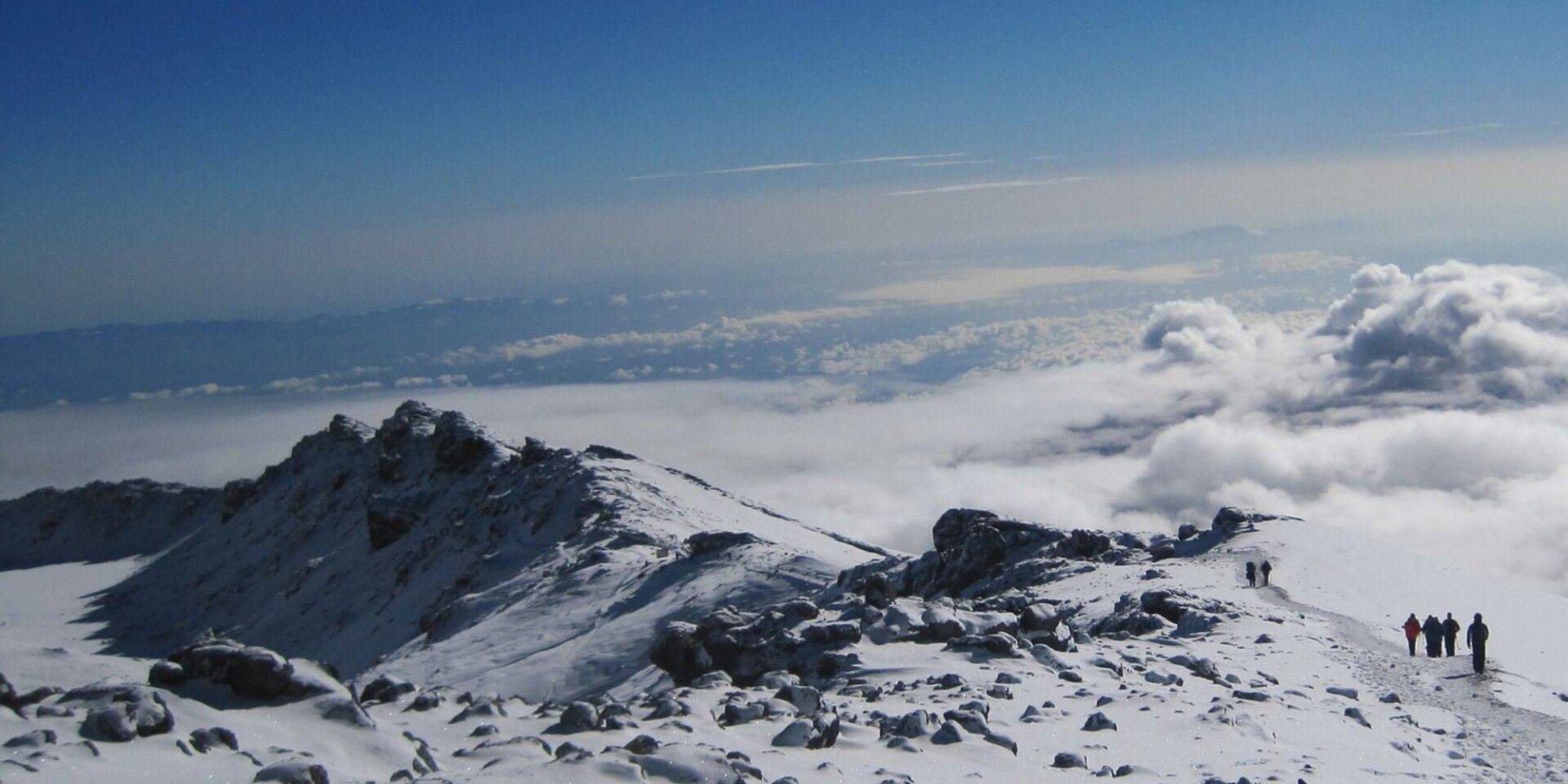 photograph of mount Kilimanjaro's summit with clouds and snow and blue sky, there are people walking to the peak