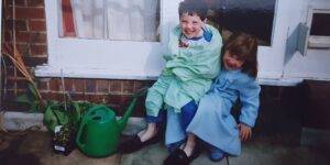 Two children sitting on a doorstep wrapped in a blanket and smiling towards the camera